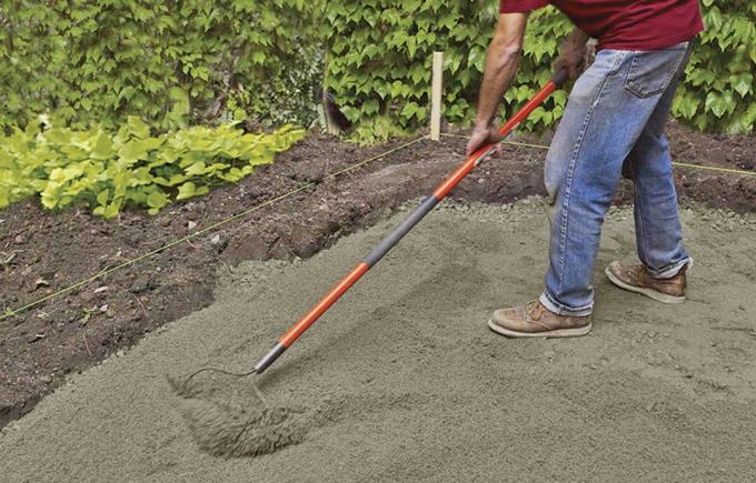 मैन ब्लूस्टोन आंगन के लिए बेस पर रेत को चिकना करने के लिए गार्डन रेक का उपयोग करता है